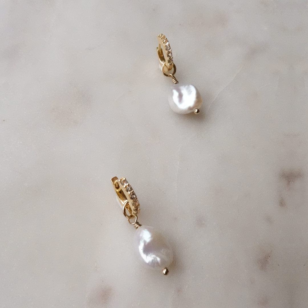 Gold filled huggies with white topaz stones and a freshwater pearl sit against a marble background.